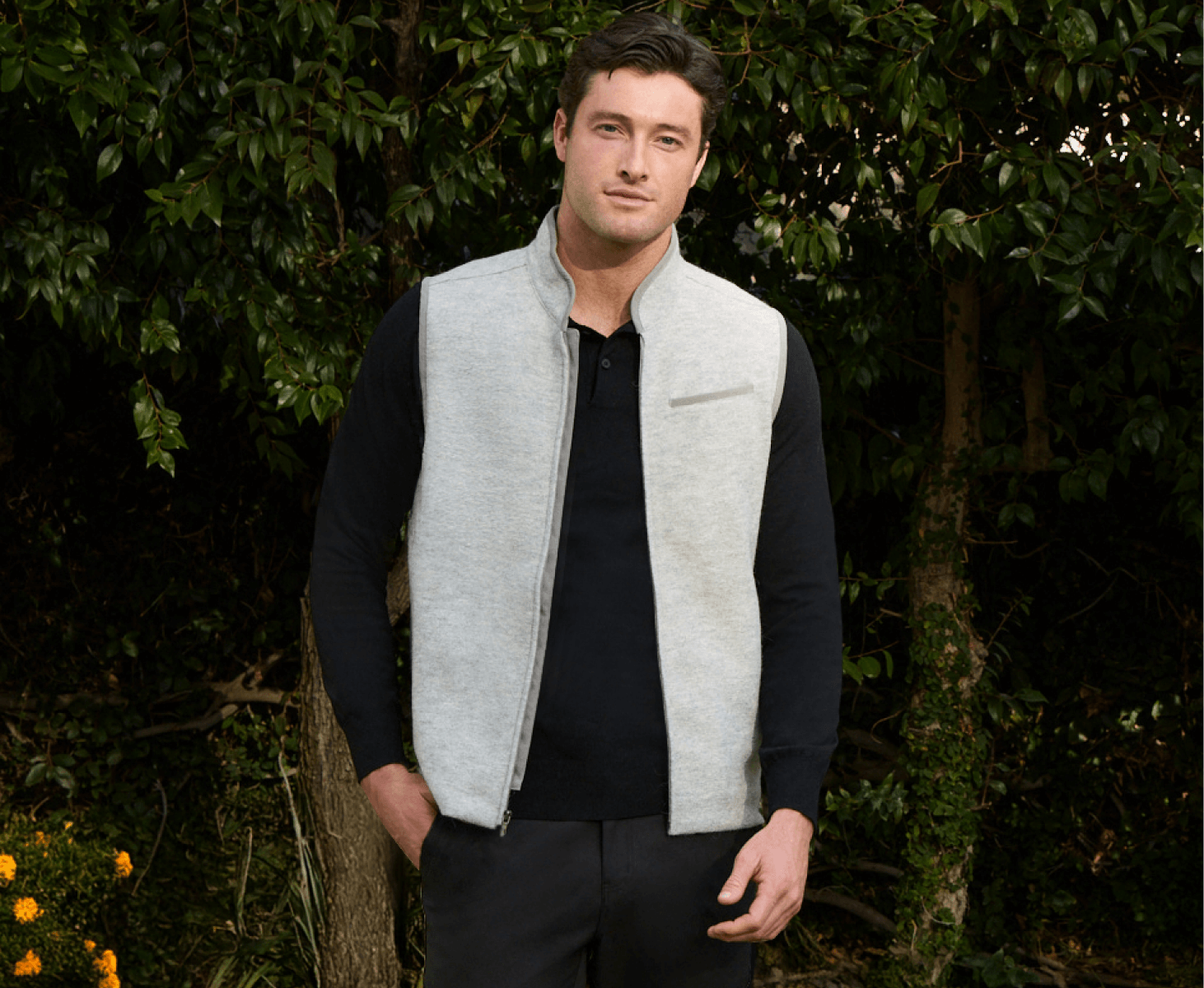 Man in a gray vest and black shirt standing outdoors with greenery in the background.