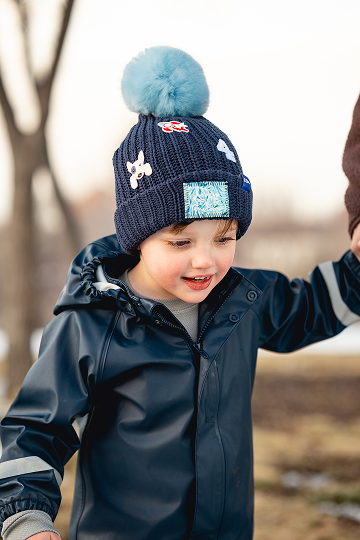 A smiling child wearing a dark blue raincoat and a matching knit hat with a light blue pom-pom.