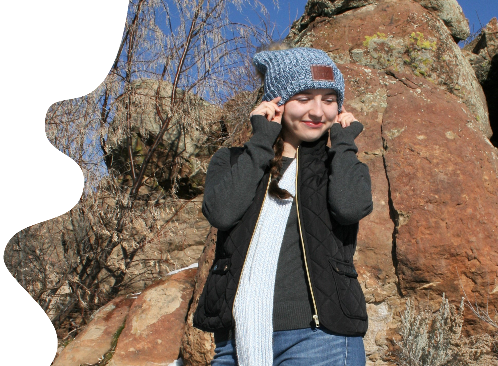 A smiling woman in a blue knit beanie adjusts her hat while standing in a rocky outdoor setting.