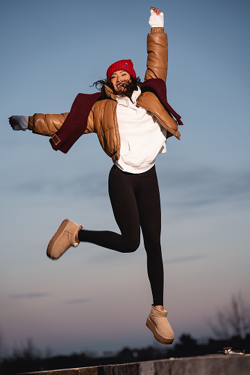 A woman in a red beanie and brown puffer jacket jumps joyfully in the air against the sky.