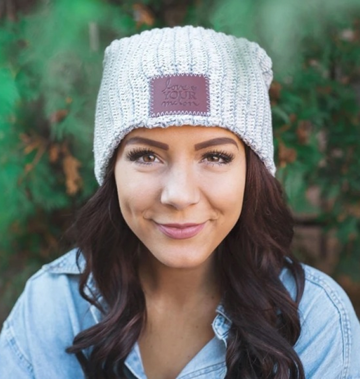 A smiling woman wears a light gray knit beanie with a 'Love Your Melon' patch and a denim shirt.