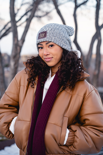 A woman in a grey knit beanie, brown puffer jacket, and maroon scarf smiles at the camera.