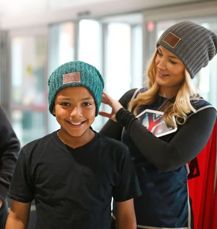 A woman in a superhero costume places a beanie on a smiling young boy's head.