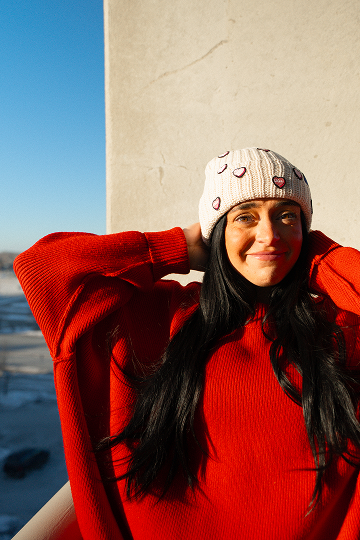 A woman in a red sweater and a cream-colored beanie with heart patches smiles at the camera.