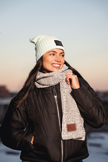 A smiling woman wearing a black puffer jacket, a light green beanie, and a grey knit scarf.