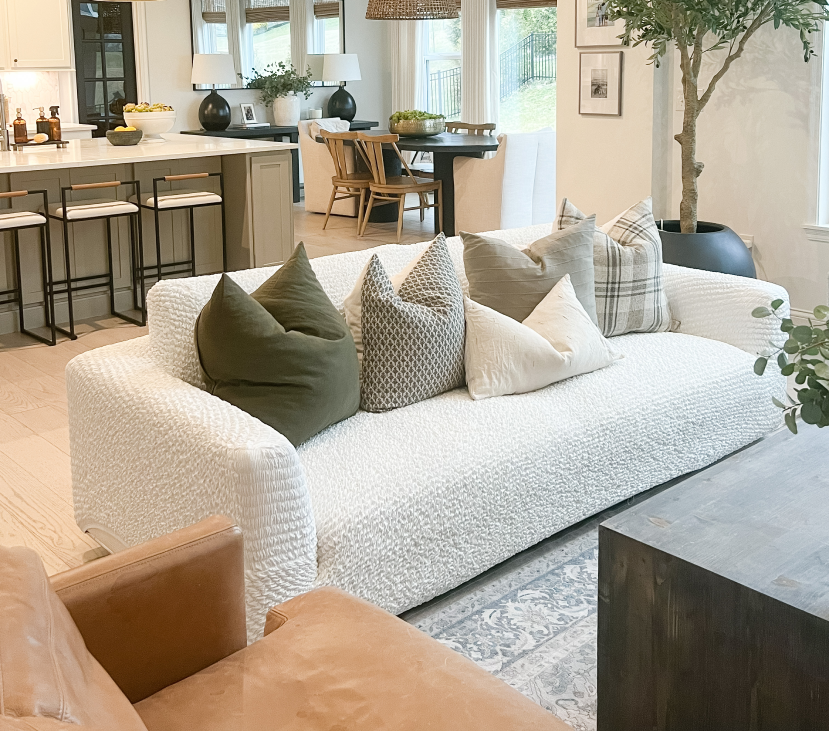 Modern living room with a white sofa, decorative pillows, and a kitchen in the background.