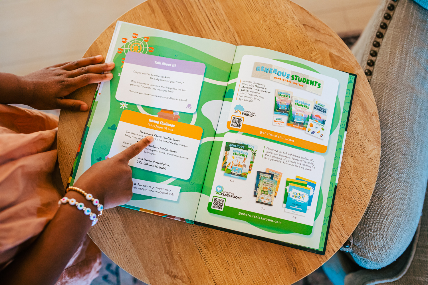 A child's hands point to a page in a colorful open book on a wooden table.