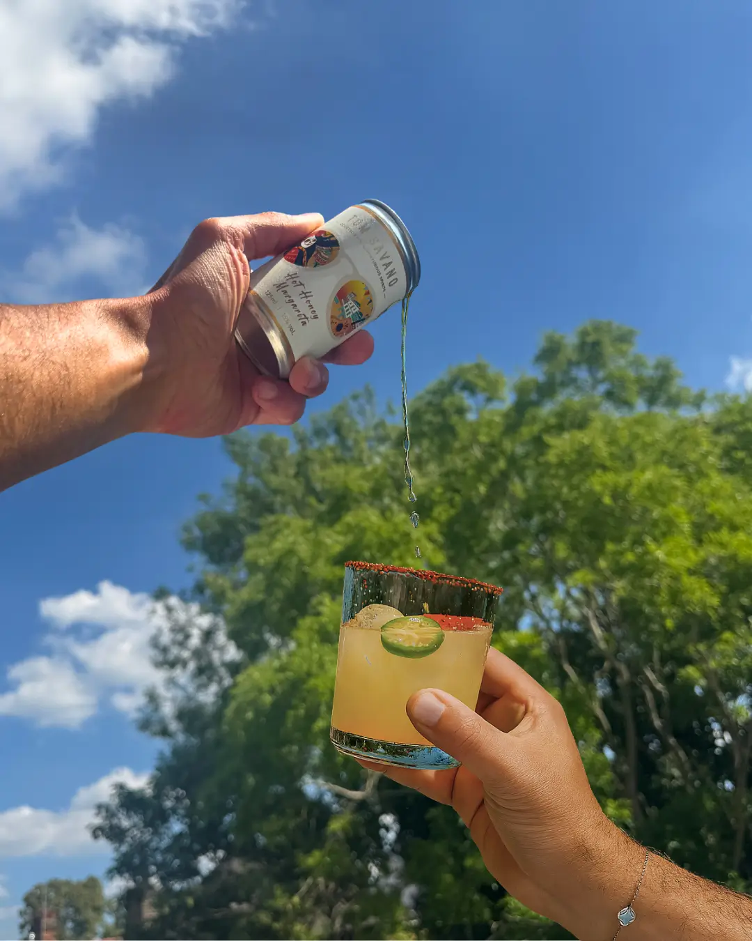 Hands pouring canned drink into a glass with lime and chili rim outdoors.