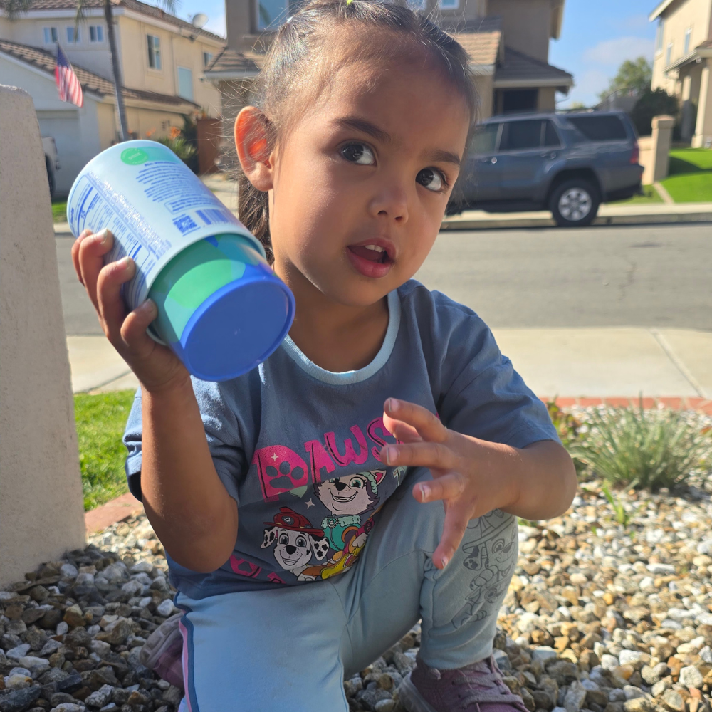 Child holding a container, sitting on gravel in a suburban neighborhood.