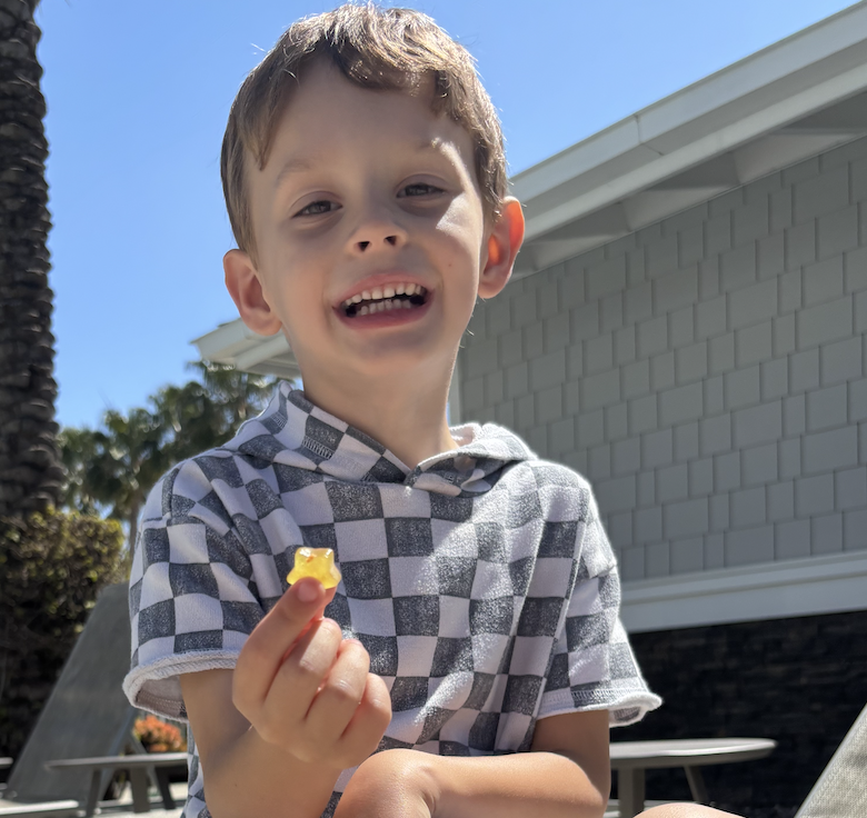 Child smiling, holding a small yellow object outdoors.