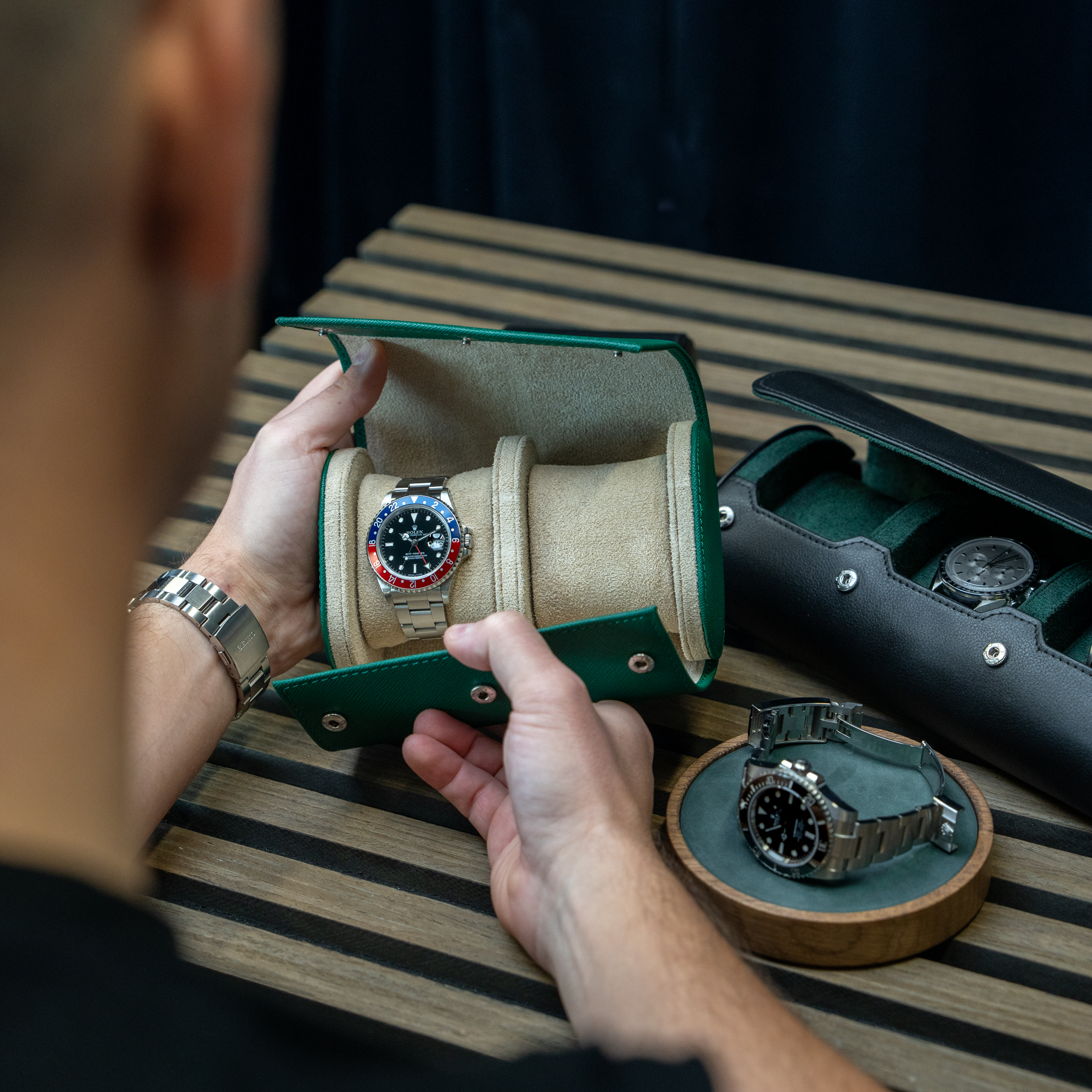A person seen from over the shoulder holds open a green leather watch roll displaying a luxury watch.