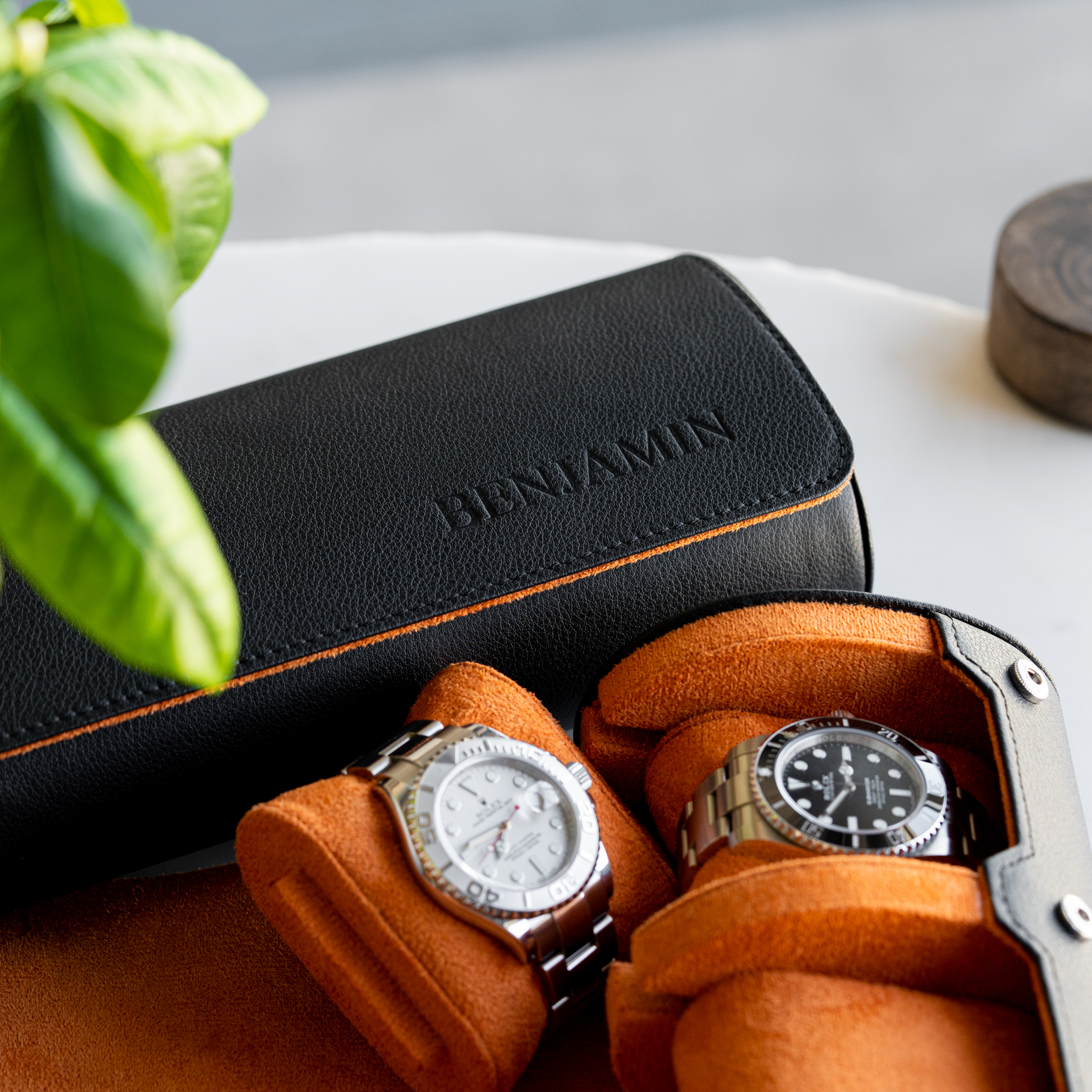 Two luxury watches sit in an open black leather travel case with a bright orange interior.