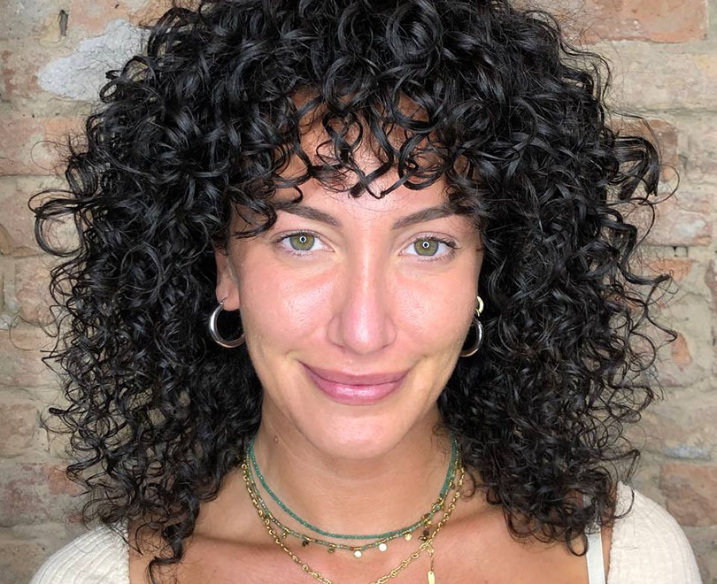Person with curly hair and hoop earrings smiling against a brick wall.