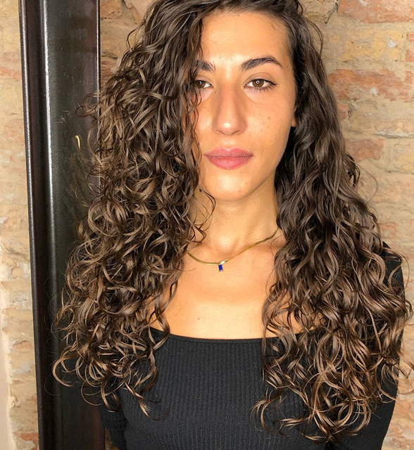 Person with long curly hair and a black top against a brick wall.