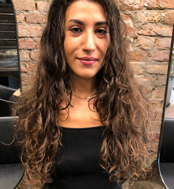 Person with long curly hair standing against a brick wall.