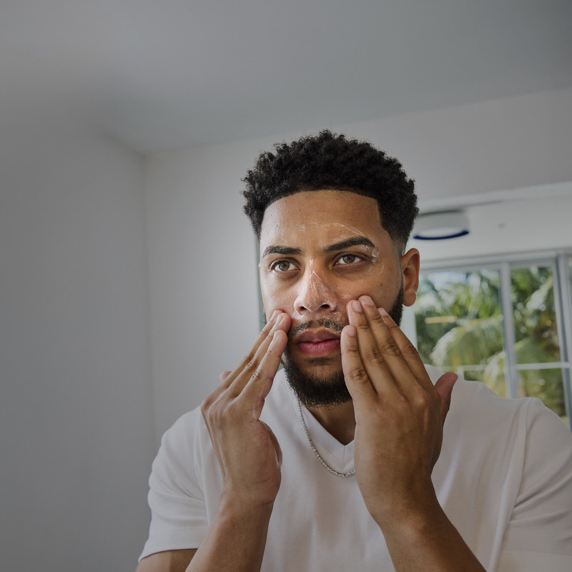 Man applying skincare product to his face in a bright room.