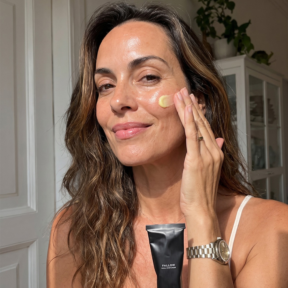 A smiling woman with glowing skin applies a yellow facial cream to her cheek while holding the product.