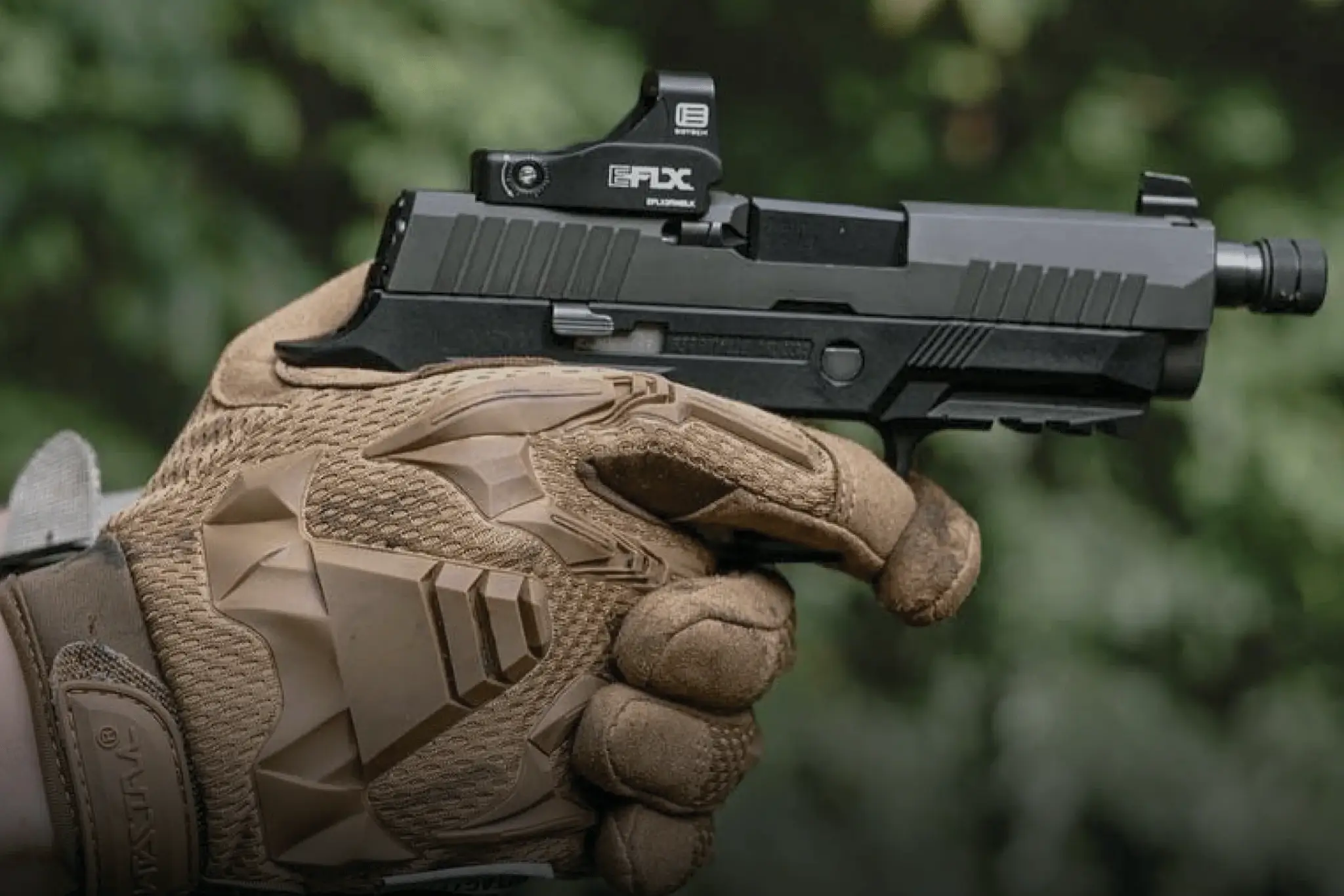 Gloved hand holding a black handgun with reflex sight.