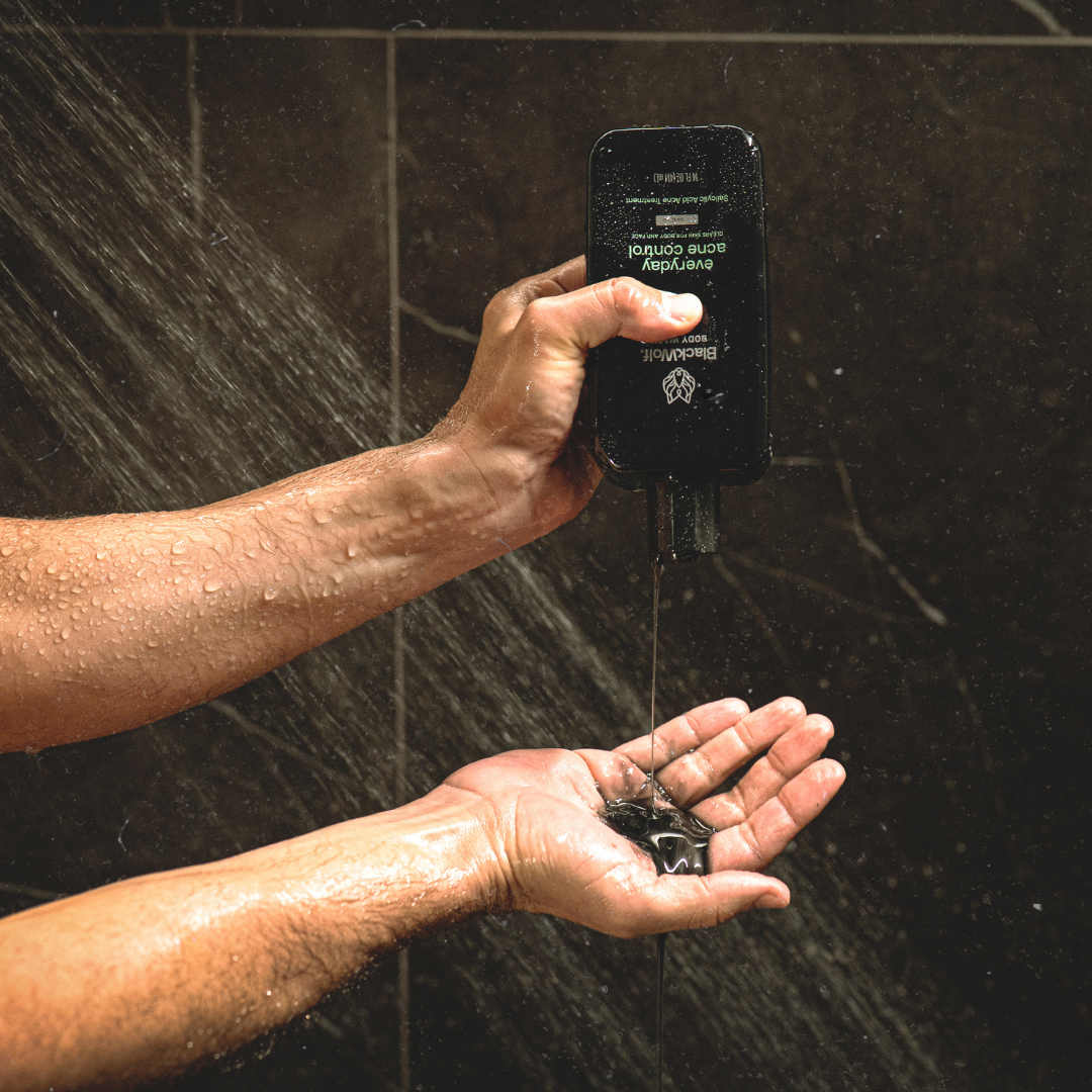 A person in a shower squeezes black body wash from a bottle into their hand.