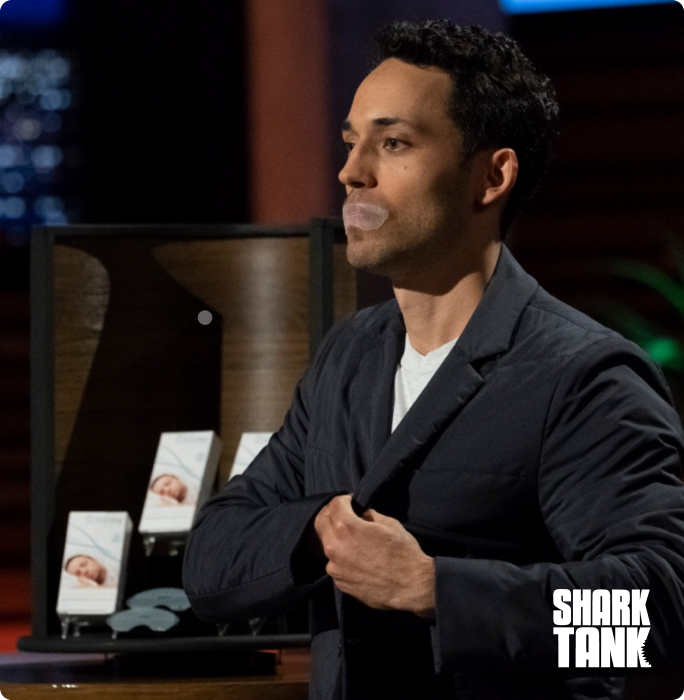 Person with tape over mouth, standing in front of product display on 'Shark Tank' set.