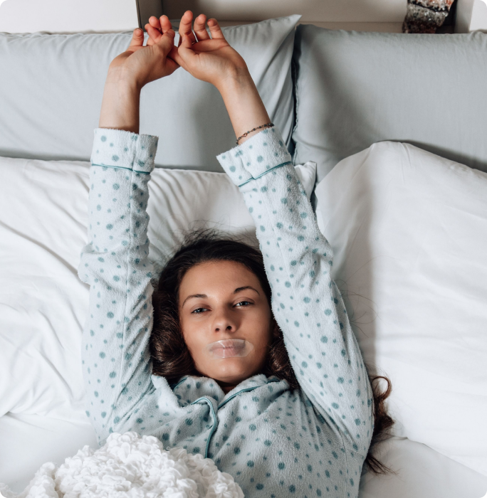 Person in polka dot pajamas stretching in bed with tape on mouth.