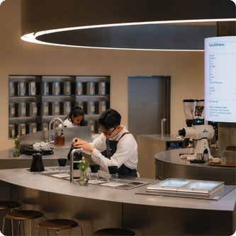 A barista in an apron prepares a drink behind a curved, modern metal counter in a coffee shop.