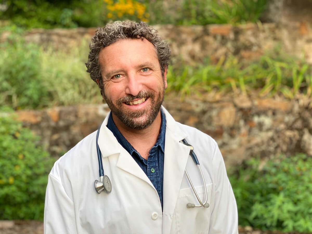 A smiling man with curly hair wearing a white doctor's coat and a stethoscope outdoors.