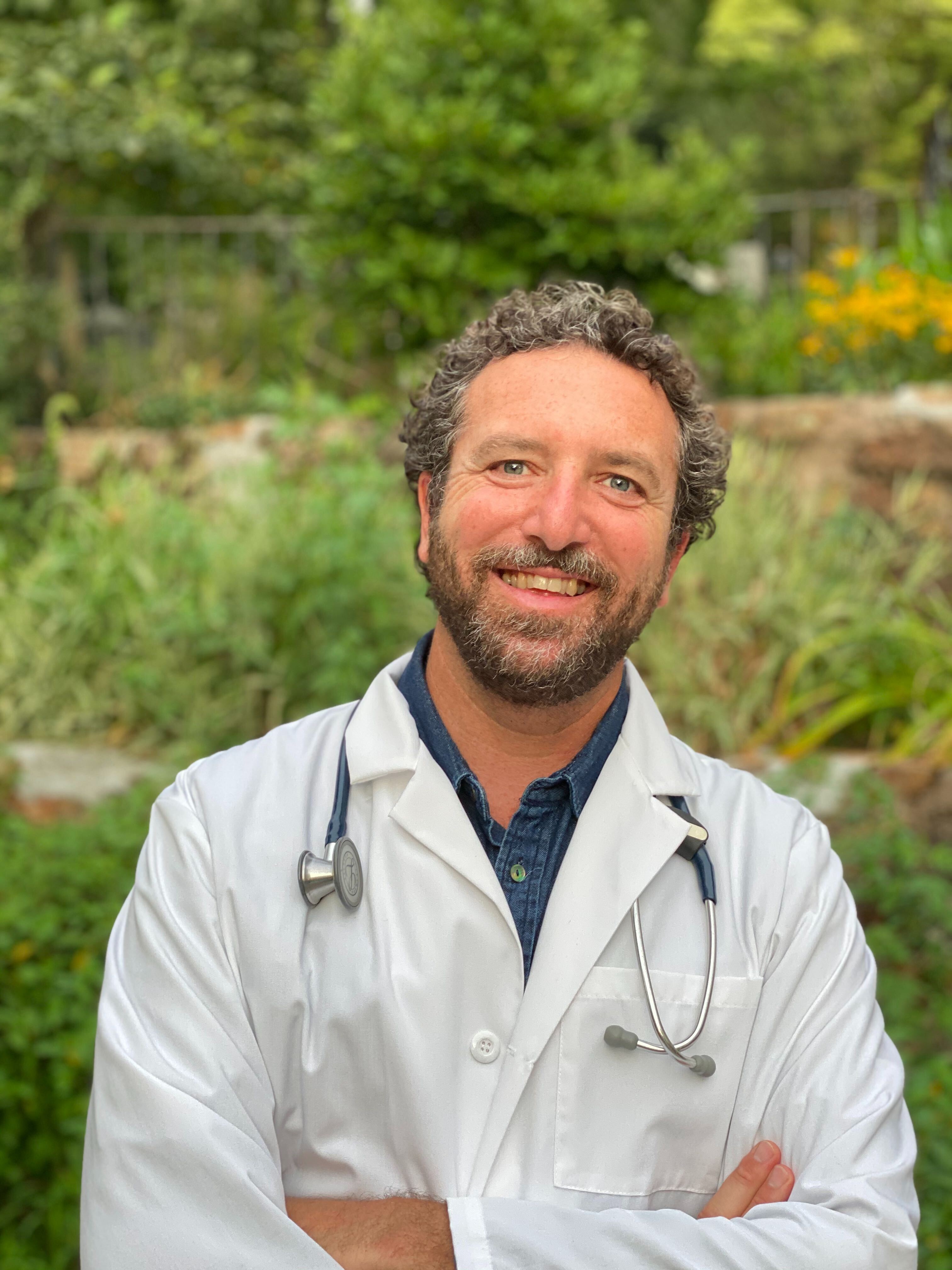 A smiling man with a beard and curly hair, wearing a white lab coat with a stethoscope outdoors.