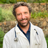 A smiling man with curly hair and a beard, wearing a white lab coat and a stethoscope.