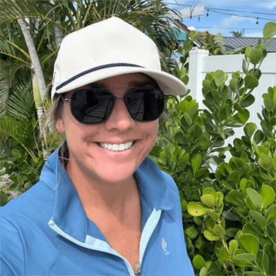 A smiling woman with grey hair and a blue collared shirt, pictured outdoors in front of green foliage.