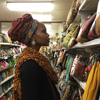 Person in a fabric store, wearing a colorful headwrap and scarf, looking at shelves of textiles.