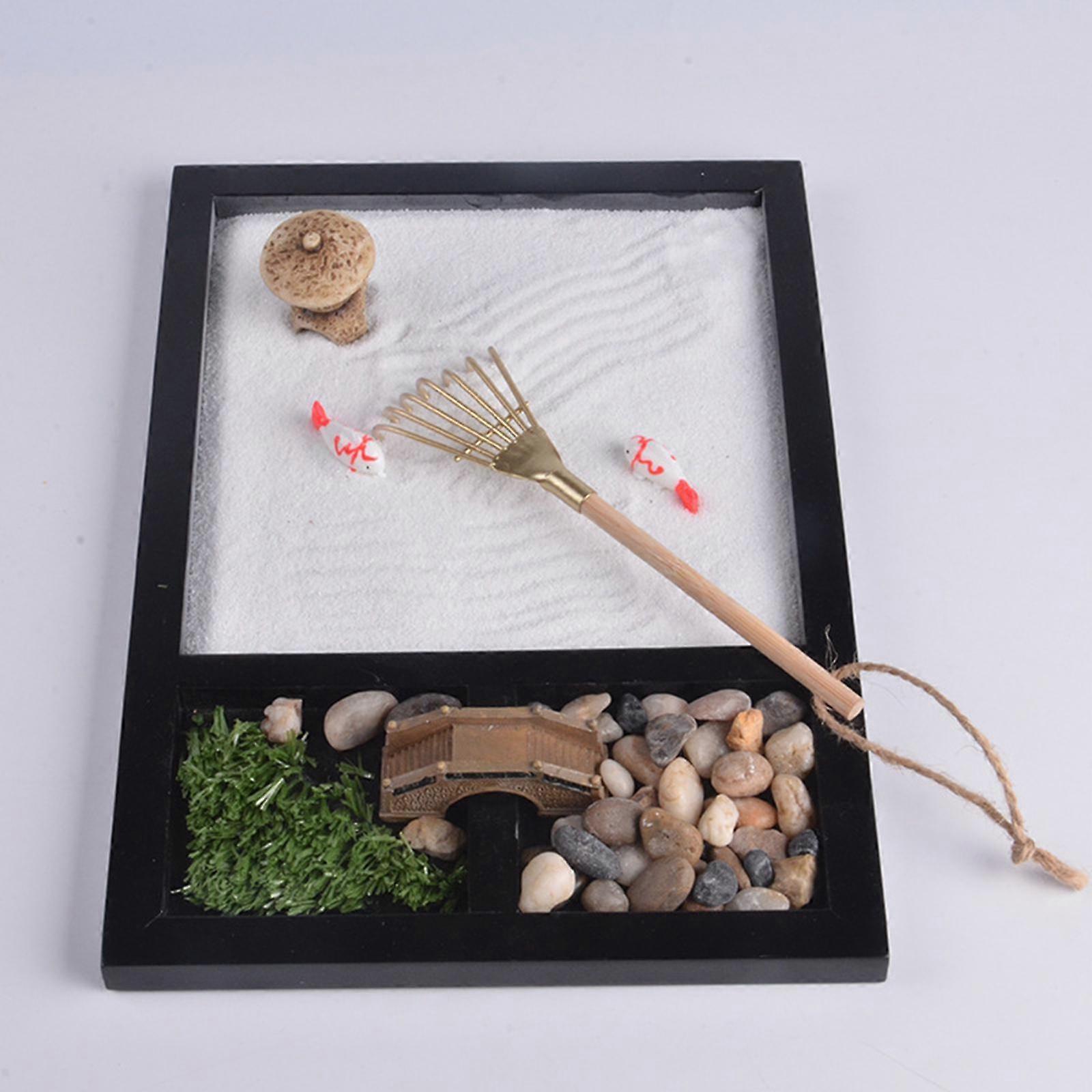 Miniature zen garden with sand, stones, a rake, and a small bridge.