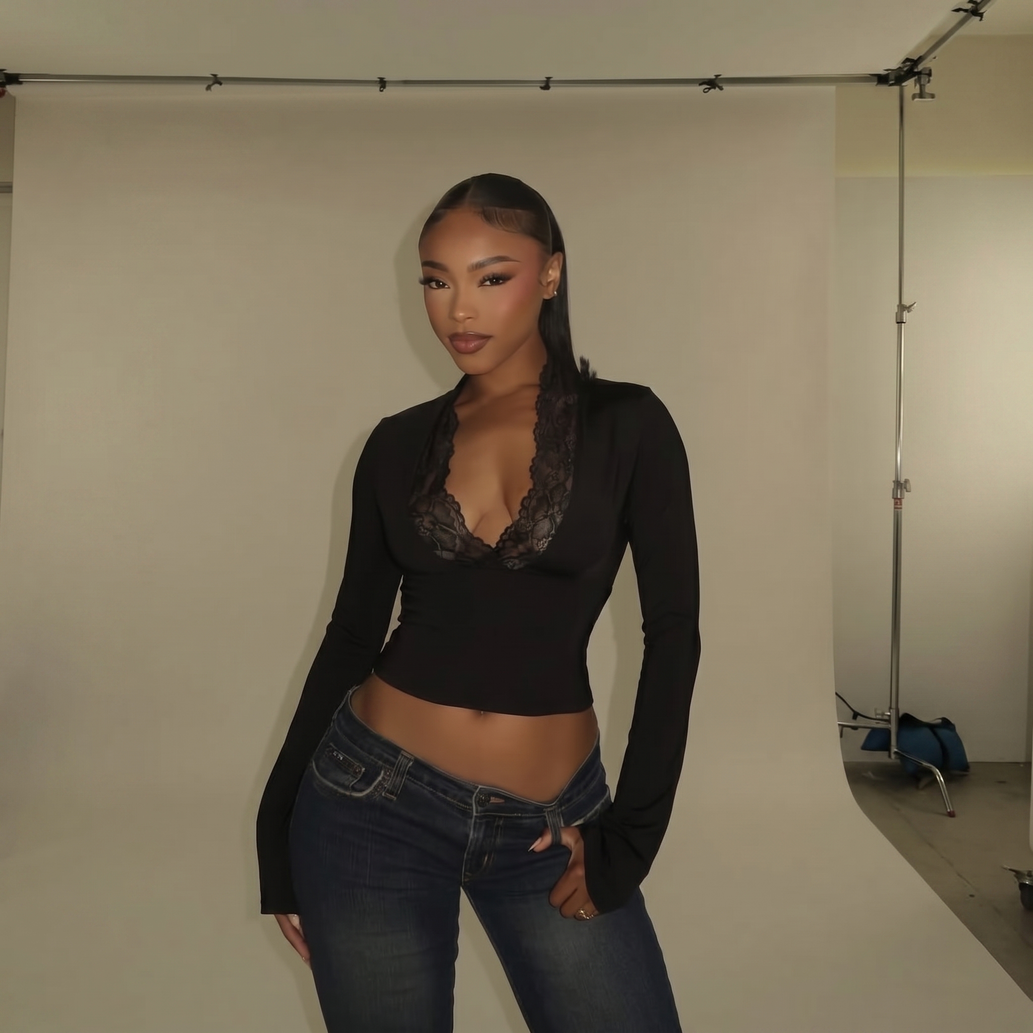 A woman poses in a photo studio wearing a black long-sleeved top and low-rise jeans.