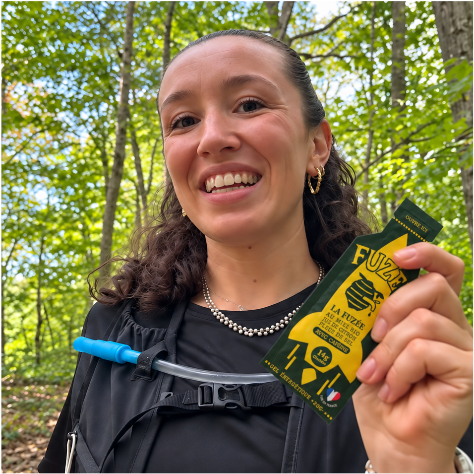 A smiling woman in a forest holds up a Fuzée energy gel packet while wearing a hydration pack.