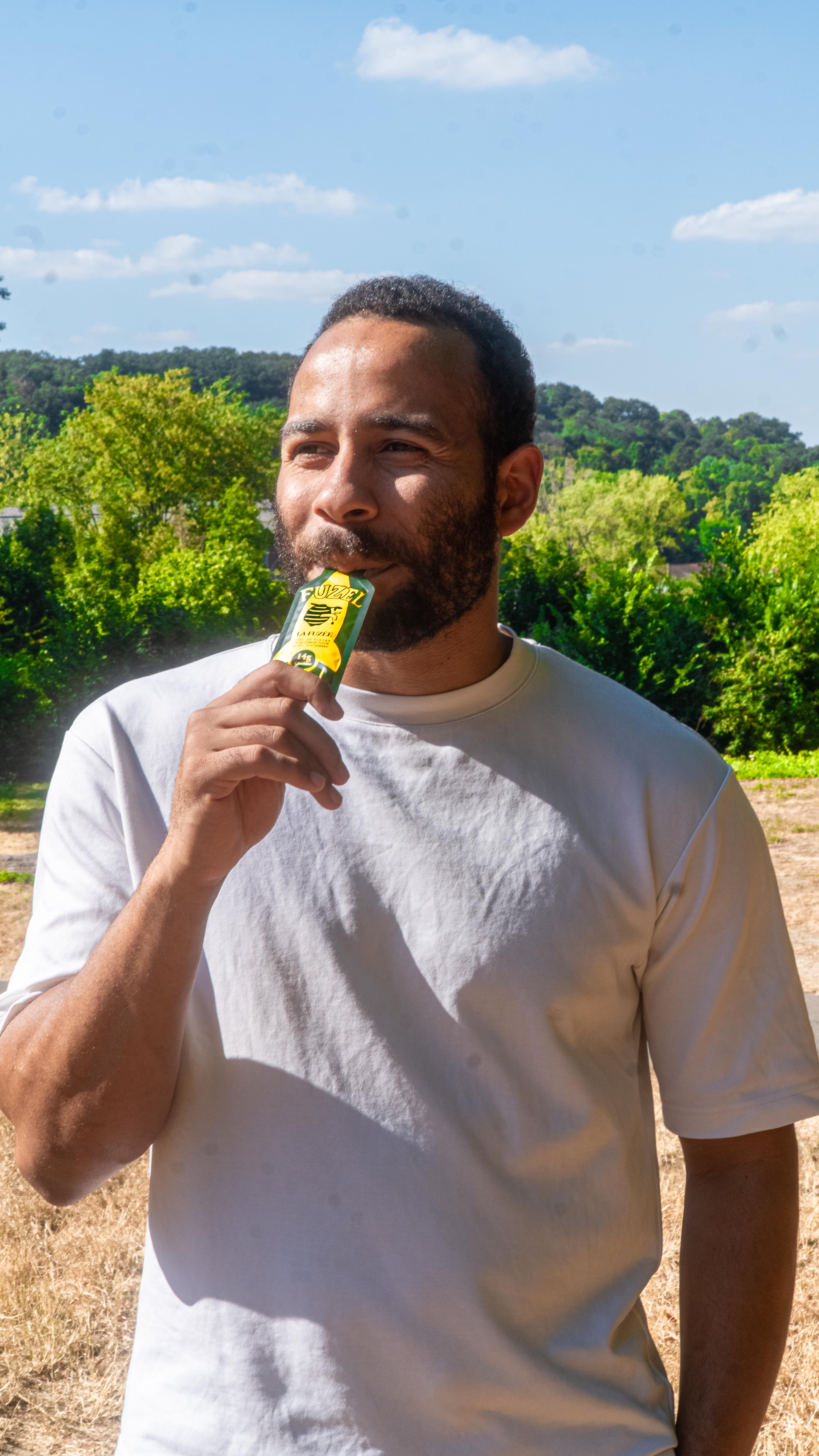 A man with a beard in a white t-shirt eats from a small packet outdoors.