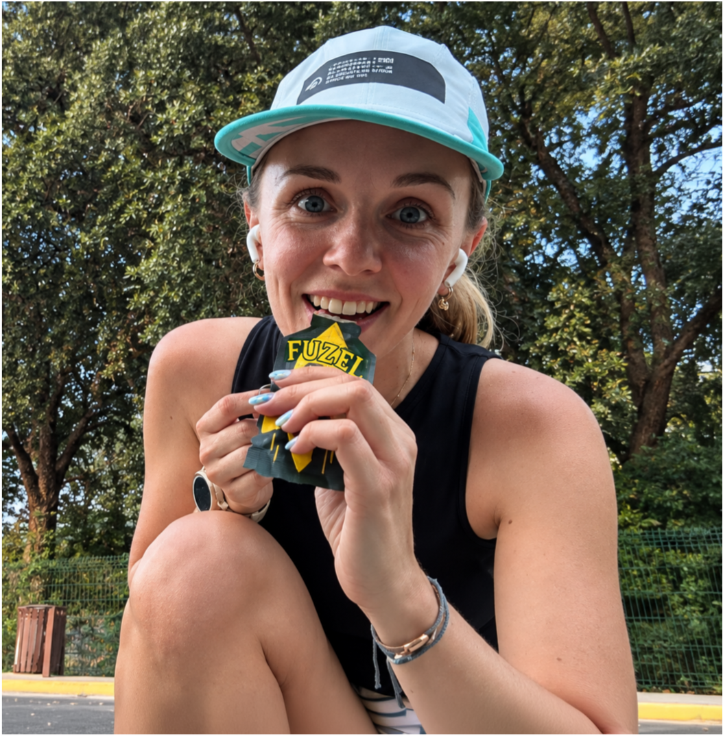 A smiling woman in a cap and earbuds eats from a FUZEL energy gel packet outdoors.