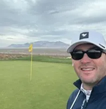 Person in sunglasses taking a selfie on a golf course with mountains in the background.