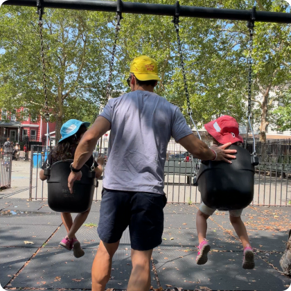 A man, seen from behind, pushes two young children on swings at an outdoor playground.