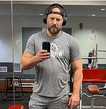 Man taking a mirror selfie in a gym, wearing headphones and a gray T-shirt.