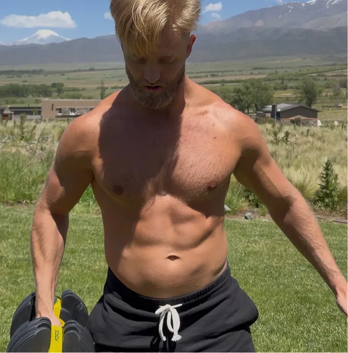 A shirtless, muscular man with a beard exercises with a dumbbell outdoors on a grassy lawn with mountains behind him.