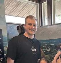 Smiling person in a black t-shirt standing indoors with posters in the background.