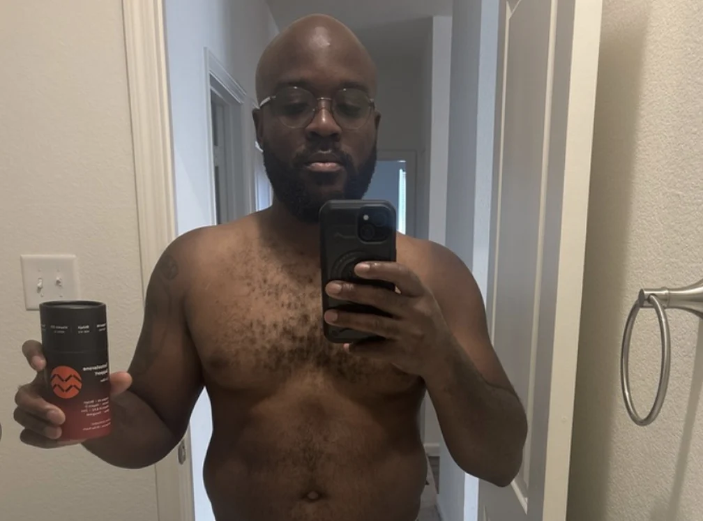 A shirtless man with glasses and a beard takes a mirror selfie while holding a product.