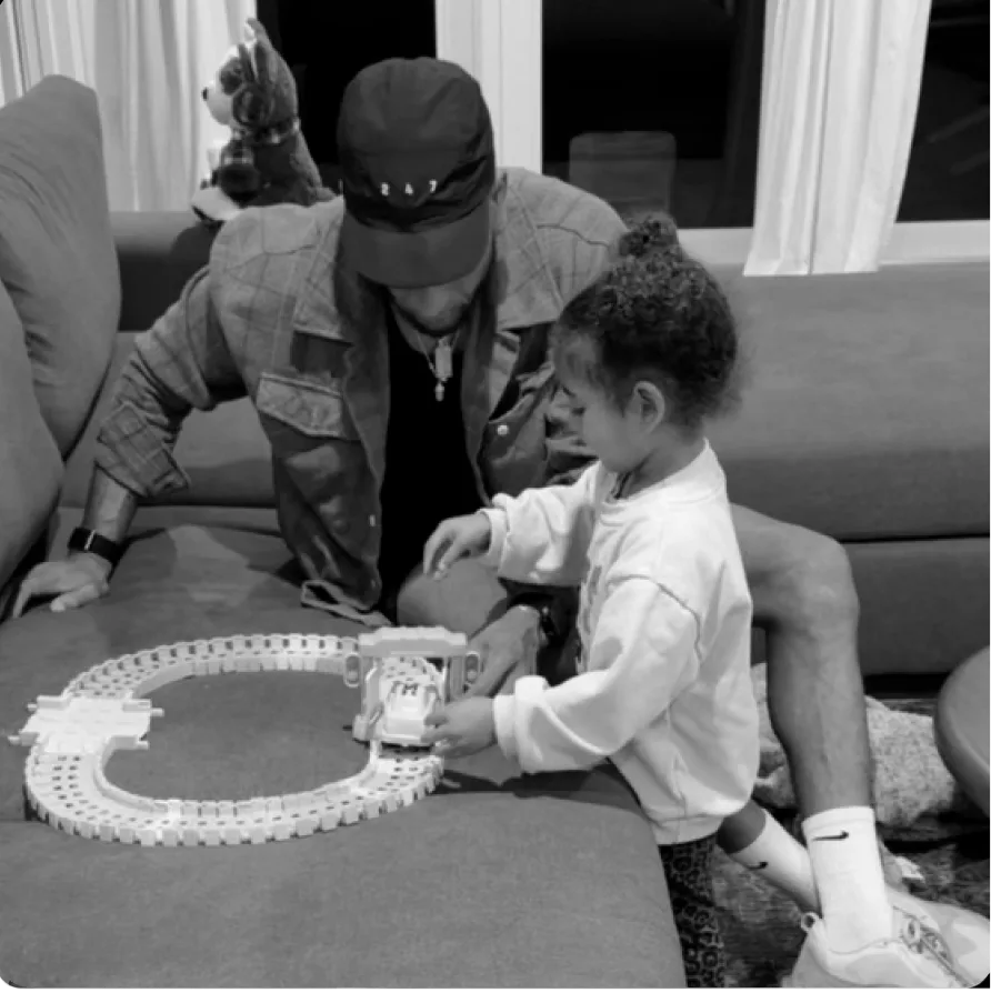 A black and white photo of a man and a small child playing with a toy train set on a couch.