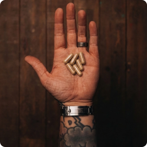 Open hand showing six brown capsules, tattooed wrist, wooden background.