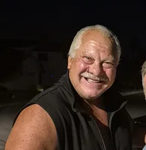 Smiling man wearing a sleeveless black jacket at night.