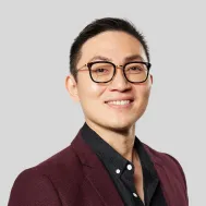 A headshot of a smiling man with glasses, wearing a black shirt and a maroon blazer.