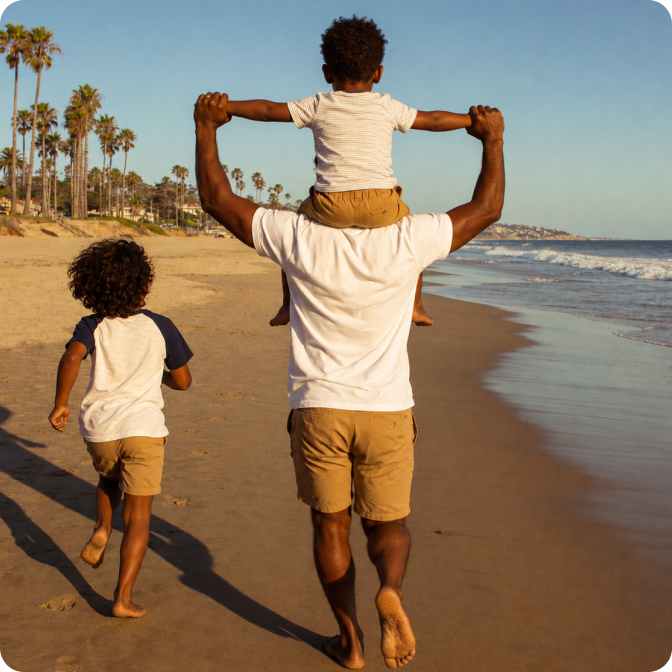 A man walks on the beach with two young boys, one of whom is riding on his shoulders.