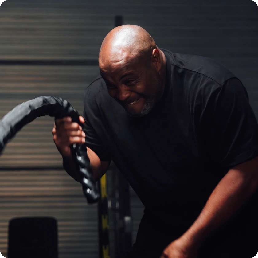 A bald man in a black shirt smiles while working out with battle ropes in a gym.