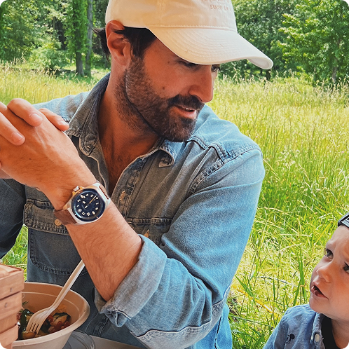 A man in a white cap and denim shirt sits in a grassy field talking to a young child.