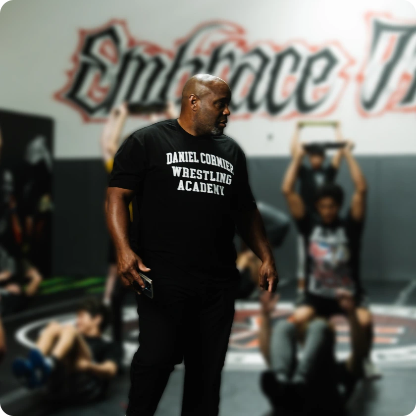 Coach Daniel Cormier, in a black t-shirt, observes students training in a wrestling academy.